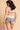 Jeune femme brune aux cheveux ondulés, de dos portant un ensemble de lingerie bleu à motifs géométrique. Elle a à la main un joli bouquet de fleurs.