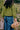 A woman with dark hair with her back to the camera. She is standing in an allotment, holding a watering can and garden trug. She wears Saywood's lambswool Adams fishermans rib knitted sweater with leaf sleeve detail, and the indigo Japanese denim Amelia Wide Leg Trousers, with a denim apron tied round her waist.