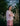 A woman stands in front of white flowering brambles, standing sideways to the camera, with her arms folded looking towards us. She wears Saywood's Amelia Wide Leg Trousers in pink corduroy with the Lela Colourblock Shirt in pink and orange. Her hair is in plaited pigtails.