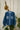 Saywood's Lela Patchwork Japanese Denim Shirt hangs on a rose gold hanger in front of a white brick wall on a rope. Behind it is a cheese plant and dried yellow flowers hanging on the wall.