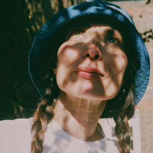 Close up of a woman standing in dappled light, looking up, with plaited pigtails. She wears Saywood's Japanese denim Reversible Bucket Hat and white recycled cotton gathered neck Marie Blouse.