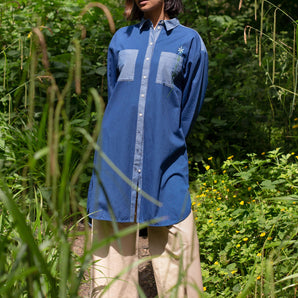 Model stands surrounded by green nature, wearing the Etta Oversized Japanese Denim Shirtdress, woven over tan trousers, with a blue bandana in recycled cotton. Hands clasped behind her back.