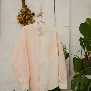 Saywood's Eddy Men's Colourblock Shirt in soft orange and pale yellow, with long sleeves, hangs on a copper hanger on a rope, in front of a white brick wall. Plants and dried flowers can be seen in the background.