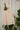 Saywood's Eddy Men's Colourblock Shirt in soft orange and pale yellow, with long sleeves, hangs on a copper hanger on a rope, in front of a white brick wall. Plants and dried flowers can be seen in the background.