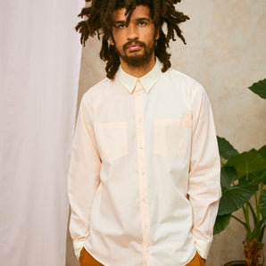 Model wears Saywood's Eddy Mens mens patchwork Shirt with patch pockets in orange and yellow. Worn with tabacco trousers. Model has both hands in his trouser pocket and a plant and drop of pink fabric can be seen in the background.