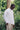 A black man wearing Saywood's Eddy Mens white Shirt with patch pockets, worn with the top buttons open and a white t-shirt underneath. He is surrounded by trees and greenery, with one hand in his trouser pocket and looking down.