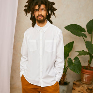 Model wears Saywood's Eddy Mens mens white Shirt with patch pockets. Worn with tabacco trousers. Model has both hands in his trouser pocket and a plant and drop of pink fabric can be seen in the background.