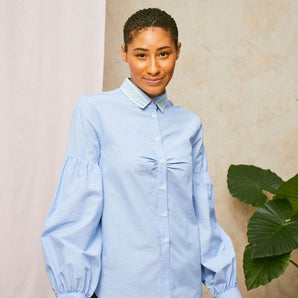 Model wears Saywood's pale blue shirt, the Edi Volume Sleeve Shirt with lace collar, with the natural indigo Japanese denim Amelia wide leg trousers. A plant and drop of pink fabric can be seen in the background. 