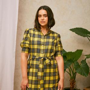 Close up crop of model wearing Saywood's Rosa yellow check shirtdress, with belt loosely tied round the waist. Puff sleeves and patch pockets with ruffles are on the dress. A plant and drop of pink fabric can be seen in the background.