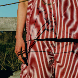 Close up of the upper body of a woman from her knees up. She has her hands by her side and is wearing Saywood's Frances Unisex Cuban Shirt and Bessie A-line Shorts in berry red stripe as a matching set. A stemmed flower is in front of her with shadows of the flowers playing on the shirt. Behind can be seen blue sky, grass and a wooden planked fence.