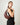 A woman wearing a black sleeveless top and black trousers carries a HYER GOODS Ring Bag large crescent-shaped red-brown leather shoulder bag with gold-tone hardware against a plain off-white background. #color_bordeaux-vintage