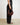 A person wearing black wide-leg trousers, a black top, and black sandals holds a HYER GOODS Rign Bag, a red-dark brown crescent-shaped leather handbag against a plain off-white wall and light floor. #color_bordeaux-vintage