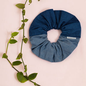 Patchwork scrunchie, made in mid wash and light wash Japanese denim. A Saywood label is stitched into the seam. The scrunchie sits on a pale pink backdrop with green foliage.