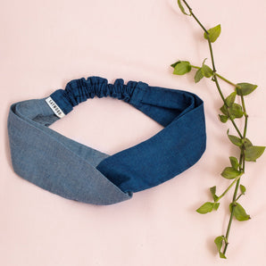 Saywood's Thandi Headband with twist effect, made from contrasting mid wash and light wash natural indigo Japanese denim. Laid on a pale pink background with green foliage.