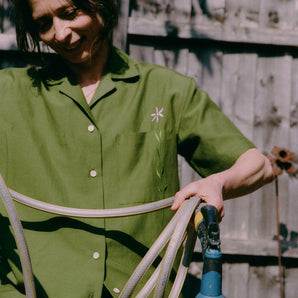 A woman is perched on a garden pot, holding a garden hose, with a wooden fence behind her. She is wearing the Frances Unisex Cuban Shirt in olive green, with short sleeves and the signature embroidered flower in lilac on the check pocket. Worn with the Bessie A-line Shorts in olive green as a matching twin set.