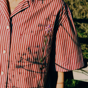 Close up of the Frances Unisex Cuban Shirt in red and white stripe. A woman is wearing the short sleeve shirt and the crop of the image shows the Saywood signature embroidered flower on the right chest pocket. Grass and a wooden fence can be seen behind her.