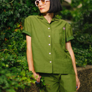 Woman wears the Saywood Frances Unisex Cuban Shirt, in olive green, with matching Bessie Aline Shorts. Made in London, the shirt is unisex, with short sleeve in a Cuban style, with an embroidered flower through the chest pocket. She wears the matching Bessie Shorts with pale pink sunglasses, and leans against a stone wall with green ivy behind her.