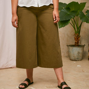 Cropped image of models legs. She is wearing Saywood's Amelia khaki wide leg trouser, cropped above the ankle, worn with black sandals. On top she wears the Joni white blouse with scalloped hem detail. A plant and drop of pink fabric can be seen in the background.