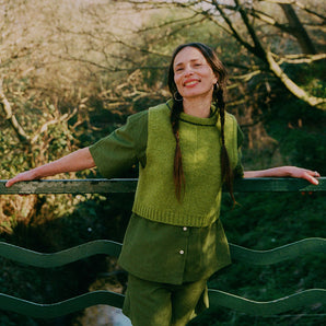 A woman with plaited pigtail hair is standing in front of green railings on a bridge overlooking trees and a small river. She is leaning away from the rails and wearing Saywood's Bessie A-line Shorts in olive green, with matching Frances Unisex Cuban Short Sleeve Shirt. Worn with the new knitted wool sweater vest from Saywood.