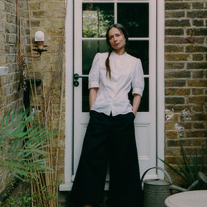 A woman stands in front of a back door in white with glass window panes. She is standing in a garden side alley, with plants in front of her and a small table. Her hands are in her trouser pockets. She wears Saywood's Amelia Wide Leg Culotte Trousers in black organic cotton and linen, with the Joni Puff Sleeve Blouse in white Supima cotton and bamboo viscose, with black open shoes.