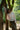 A woman stands, with one arm up leaning against the trunk of a tree. Trees and greenery can be seen behind her. She is wearing Saywood's Lela Colourblock Boxy shirt in lemon and orange vibrant pastel, with the Amelia Wide Leg Trousers in khaki. She has sunglasses on.