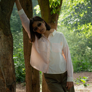 A woman stands, with one arm up leaning against the trunk of a tree. Trees and greenery can be seen behind her. She is wearing Saywood's Lela Colourblock Boxy shirt in lemon and orange vibrant pastel, with the Amelia Wide Leg Trousers in khaki. She has sunglasses on.