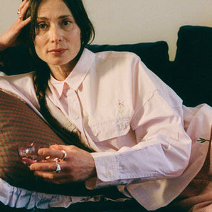 Close up image of a woman lounging on a navy sofa, leaning on a Saywood red check and lilac ruffle cushion. She is wearing Saywood's lilac and pink colourblock Jules Utility Shirt with the pink corduroy Amelia Trousers. The embroidered flower is visible on the check of the shirt above the pocket, and at the hem. She is holding a pink glass of wine with chunky silver rings on her fingers. 