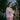 A woman stands in front of white flowering brambles, standing sideways to the camera, with her arms folded looking towards us. She wears Saywood's Amelia Wide Leg Trousers in pink corduroy with the Lela Colourblock Shirt in pink and orange. Her hair is in plaited pigtails.