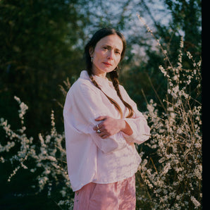 A woman stands in front of white flowering brambles, standing sideways to the camera, with her arms folded looking towards us. She wears Saywood's Amelia Wide Leg Trousers in pink corduroy with the Lela Colourblock Shirt in pink and orange. Her hair is in plaited pigtails.