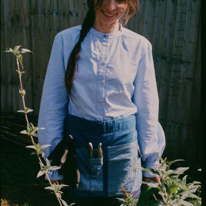 A woman stands smiling in her garden, looking at plants. She is wearing Saywood's Marie A-line Blouse with gathered neck and cuffs, with a blue vintage glass button at the neckline. Around her waist is the Multifunctional Gardening Tool Belt Apron in Japanese Denim with gardening hand tools in.