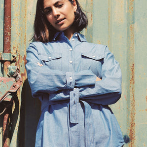 Model leans against a green shipping container, with arms crossed in front of her waist. She wears Saywood's Zadie Boyfriend Shirt in Japanese Denim with a pair of beige trousers.