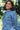 Close up of a woman standing amongst leafy trees, turned at a slight angle. She is wearing Saywood's Zadie Oversized Shirt in lightwash Japanese denim, with the tied belt tied around the waist. The utility patch pockets and top stitch detailing can be seen.