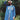 A woman stands surrounded by green nature, wearing the Etta Oversized Japanese Denim Shirtdress, worn over tan trousers, with a blue bandana in recycled cotton. One hand is held up to the sky to block the sunlight.