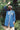 A woman stands surrounded by green nature, wearing the Etta Oversized Japanese Denim Shirtdress, worn over tan trousers, with a blue bandana in recycled cotton. One hand is held up to the sky to block the sunlight.