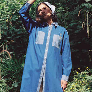 A woman stands surrounded by green nature, wearing the Etta Oversized Japanese Denim Shirtdress, worn over tan trousers, with a blue bandana in recycled cotton. One hand is held up to the sky to block the sunlight.
