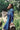 A woman sits on a log, shot from the side, surrounded by green nature, looking towards the camera. She is wearing the Etta Oversized Japanese Denim Shirtdress, worn over tan trousers.