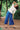 A woman stands out in nature surrounded by trees. She stands with her legs a part leaning bent to one side with one hand on her head. She wears Saywood's Amelia Wide Leg Culotte Trousers in Japanese denim with the Joni fit and flare blouse and scalloped hem in white. Worn with sandals and a red check neck tie.