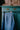 The Japanese denim Amelia Wide Leg Culotte Trousers are hanging on a rose gold hanger from the ornate gold handle of a turquoise vintage wooden cupboard, with metal cabinet on top. The back of the trousers can be seen, with the signature Saywood embroidered flower on the back pocket, and triangle cut out at the waist. The trousers have been worn so the denim has slightly washed down with age.