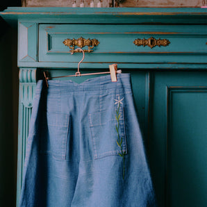 The Japanese denim Amelia Wide Leg Culotte Trousers are hanging on a rose gold hanger from the ornate gold handle of a turquoise vintage wooden cupboard, with metal cabinet on top. The back of the trousers can be seen, with the signature Saywood embroidered flower on the back pocket, and triangle cut out at the waist. The trousers have been worn so the denim has slightly washed down with age.
