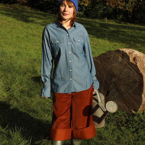 Women standing in a green field with trees behind her and a big tree trunk in the background. She is wearing Saywood's Caramel Tan Corduroy Trousers in Organic Cotton, styled rolled up with wellies, with the cotton Zadie Boyfriend Shirt in Japanese Denim, and a blue beanie hat. She holds a watering can.