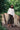 Model stands, one knee bent, in front of tree stumps and greenery. She wears Saywood's Amelia wide leg trouser in khaki cotton sateen, with the white Marie A-line Blouse with a red check neckerchief tied round her neck. Worn with black and white zebra print sandals.