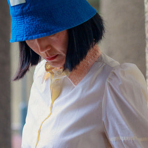Close up of a woman with dark hair, looking downwards. She is wearing Saywood's Joni Puff Sleeve Blouse in lemon, with fit and flare shaping, peach lace collar and scalloped hem. Worn with the Japanese denim Bucket Hat.