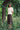 A woman stands on a tree stump, with her hands behind her back, surrounded by nature. She is wearing Saywood's Joni Puff Sleeve Blouse, with fit and flare darting, scallop hem adn lace trim on the collar. Worn with the Amelia Wide Leg Trousers in khaki cotton, sunglasses and black sandals.