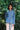 A woman stands amongst leafy trees, with one arm behind her back. She is wearing Saywood's Zadie Boyfriend Fit shirt in lightwash Japanese denim, worn with light tan chinos.