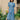 A woman stands wearing Saywood's Rosa denim shirtdress in light wash Japanese denim; a long line style with patch pockets and ruffles, a belt tied round the waist. She stands with her hands by her side at ease. Yellow flowers can be seen in the background.