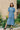 A woman stands wearing Saywood's Rosa denim shirtdress in light wash Japanese denim; a long line style with patch pockets and ruffles, a belt tied round the waist. She stands with her hands by her side at ease. Yellow flowers can be seen in the background.