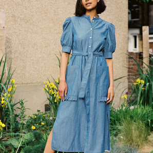 A woman stands wearing Saywood's Rosa denim shirtdress in light wash Japanese denim; a long line style with patch pockets and ruffles, a belt tied round the waist. She stands with her hands by her side at ease. Yellow flowers can be seen in the background.