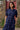 A woman stands in front of a brick wall and red wooden door. She is wearing Saywood's Rosa Puff Sleeve Shirtdress in navy check with the  belt loosely tied around her waist. The gathered puff sleeves and patch pockets with ruffle trims can be seen in the close up shot.