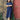 A woman leans against a brick wall with a red gate door next to her. She has her arms folded and wears Saywood's Rosa puff sleeve dress in navy check with black sandals. Plants can be seen growing out near her feet.