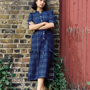 A woman leans against a brick wall with a red gate door next to her. She has her arms folded and wears Saywood's Rosa puff sleeve dress in navy check with black sandals. Plants can be seen growing out near her feet.